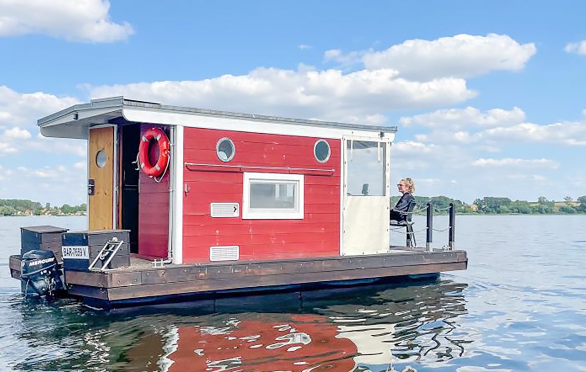 Rotes schwimmendes Floß auf dem Wasser mit Person am Steuer. Heller Himmel mit Wolken.